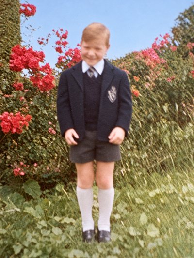 A boy in school uniform and blazer standing in front of rose bushes.