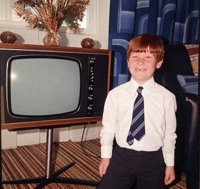 Jimi Gill as a boy in school shirt and tie, standing beside a wood-cased television.