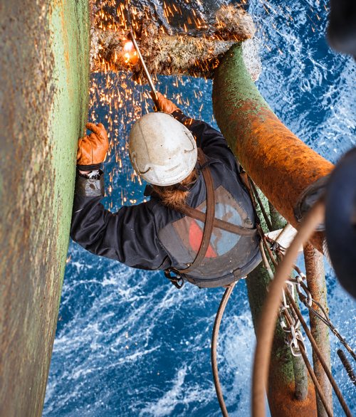 A rope access technician cutting steel high above open water, sparks falling into the sea.